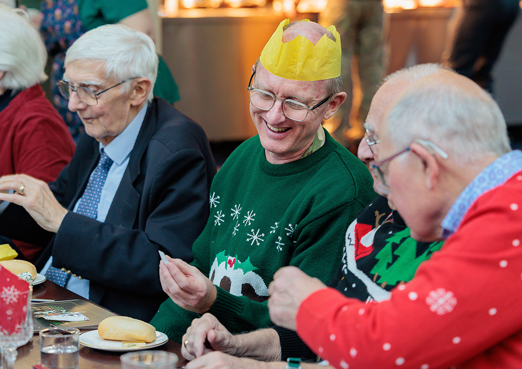 This photo shows invited Veterans from the local community, sitting at a prepared lunch table while sharing Christmas cracker jokes.